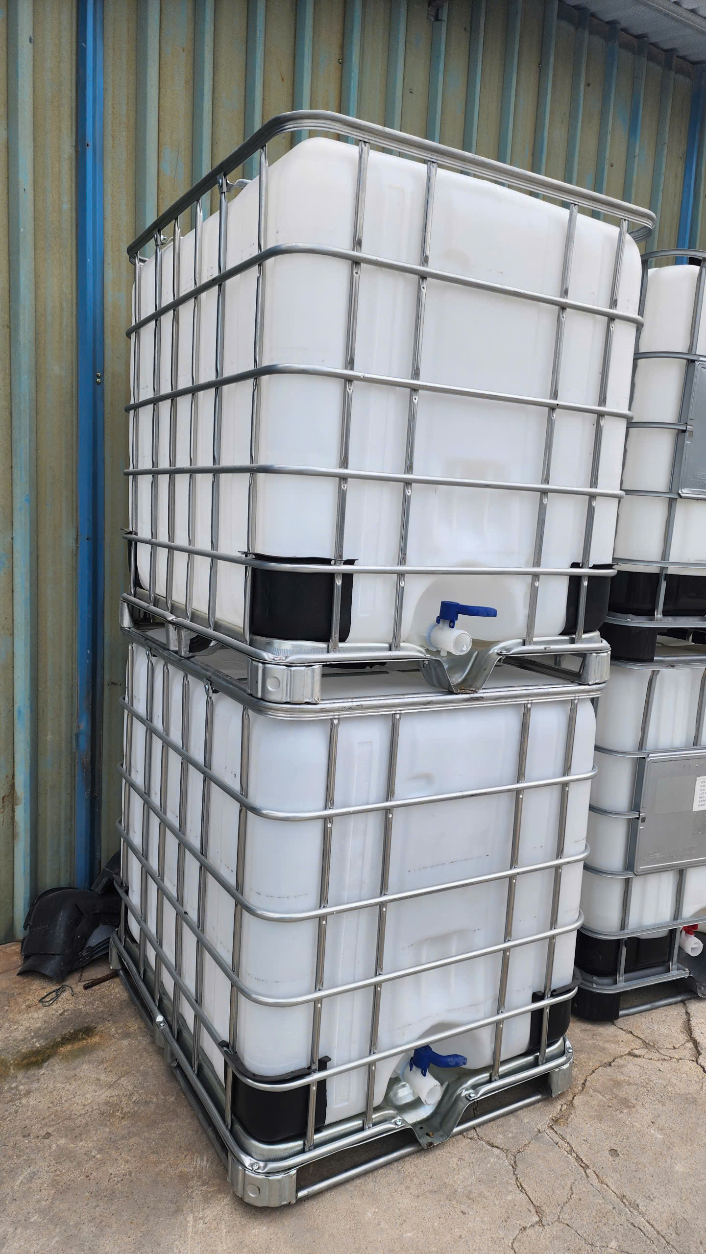 Hai tank IBC 1000L cũ đặt chồng cạnh vách tôn, mỗi tank có van cần gạt xanh ở đáy